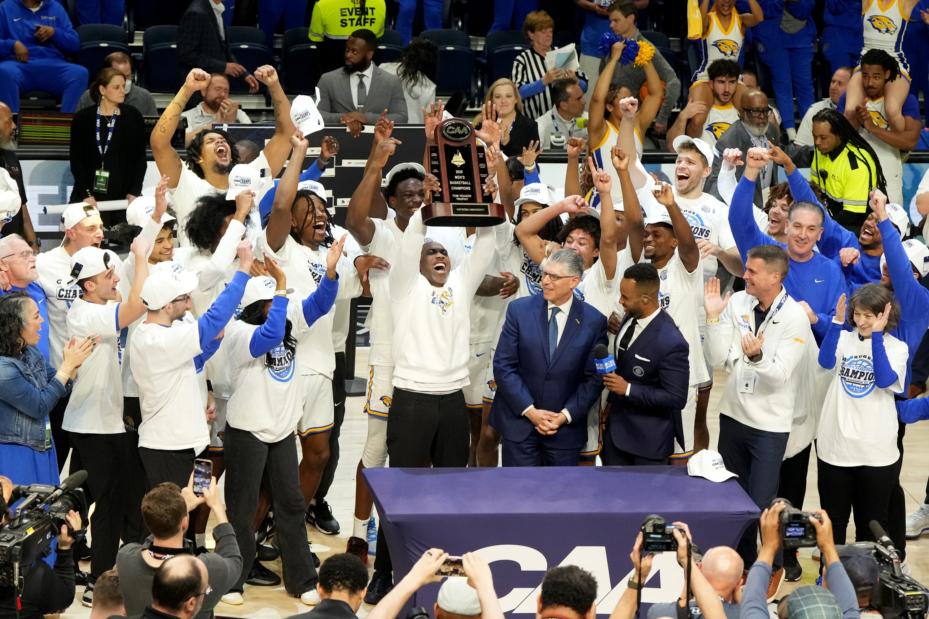 The Hofstra Pride men's basketball team celebrates their championship win, with players and staff raising a trophy in excitement.
